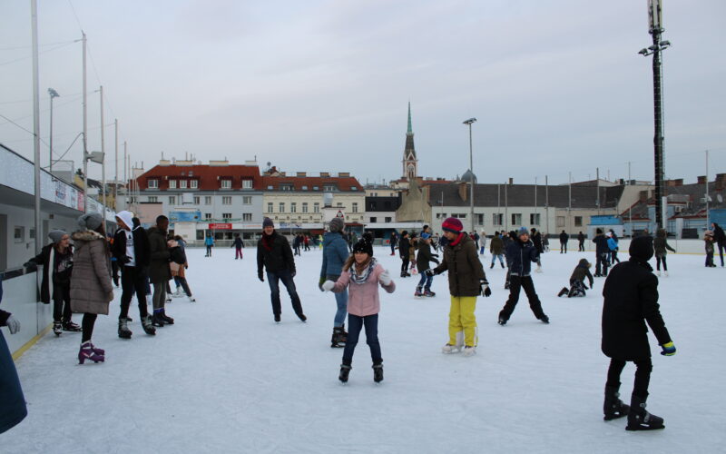 Menschen verschiedenen Alters beim Schlittschuhlaufen auf einer Freiluft-Eisbahn in einer Stadt, mit Gebäuden und einem Kirchturm im Hintergrund.