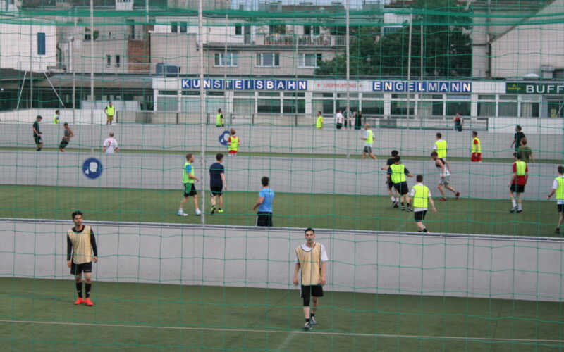 Mehrere Gruppen von Menschen spielen Fußball auf einem Kunstrasenplatz im Freien, getrennt durch Netze, mit Gebäuden im Hintergrund.