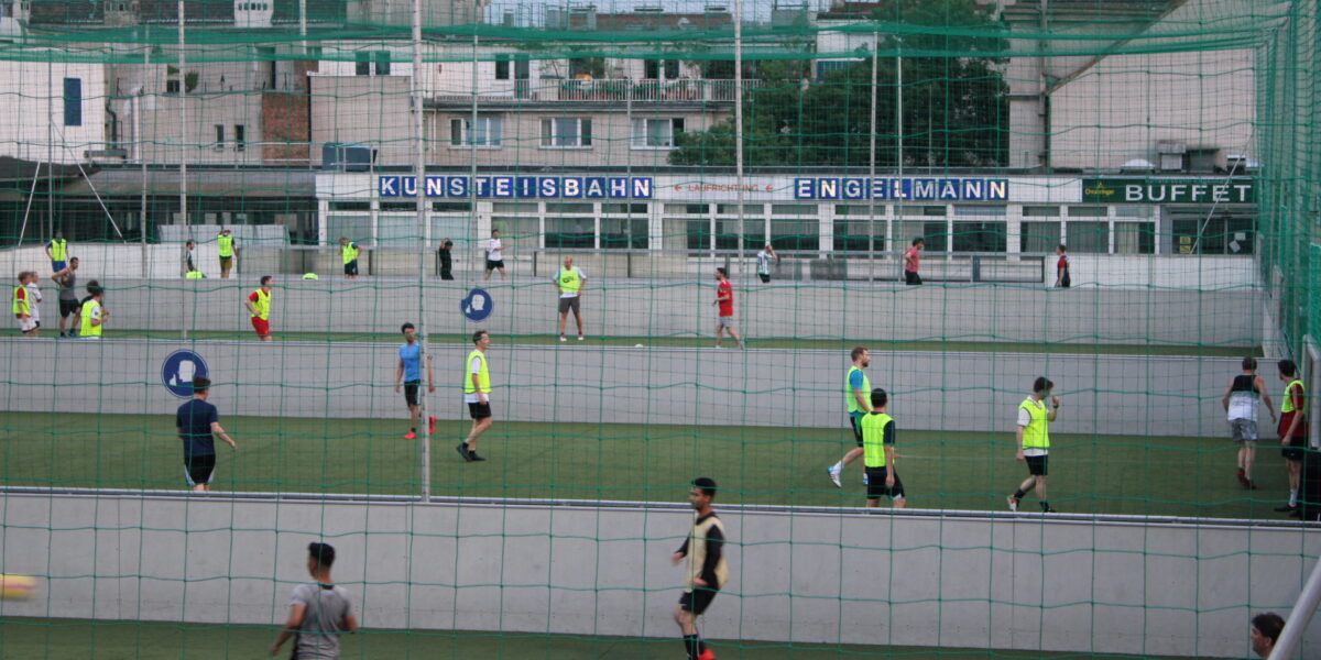Mehrere Personen spielen Fußball auf einem mit Netzen eingefassten Feld im Freien, im Hintergrund sind Gebäude und ein Schild mit der Aufschrift "KUNSTEISBAHN ENGELMANN BUFFET" zu sehen.