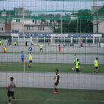 Mehrere Personen spielen Fußball auf einem mit Netzen eingefassten Feld im Freien, im Hintergrund sind Gebäude und ein Schild mit der Aufschrift "KUNSTEISBAHN ENGELMANN BUFFET" zu sehen.