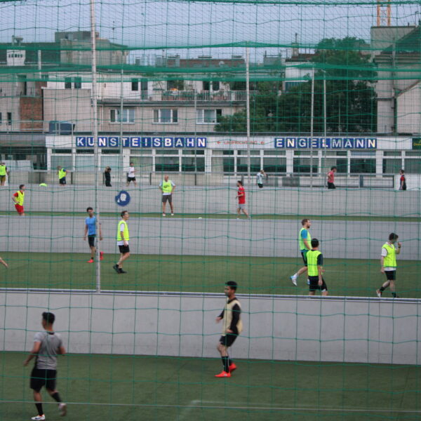 Mehrere Personen spielen Fußball auf einem mit Netzen eingefassten Feld im Freien, im Hintergrund sind Gebäude und ein Schild mit der Aufschrift "KUNSTEISBAHN ENGELMANN BUFFET" zu sehen.