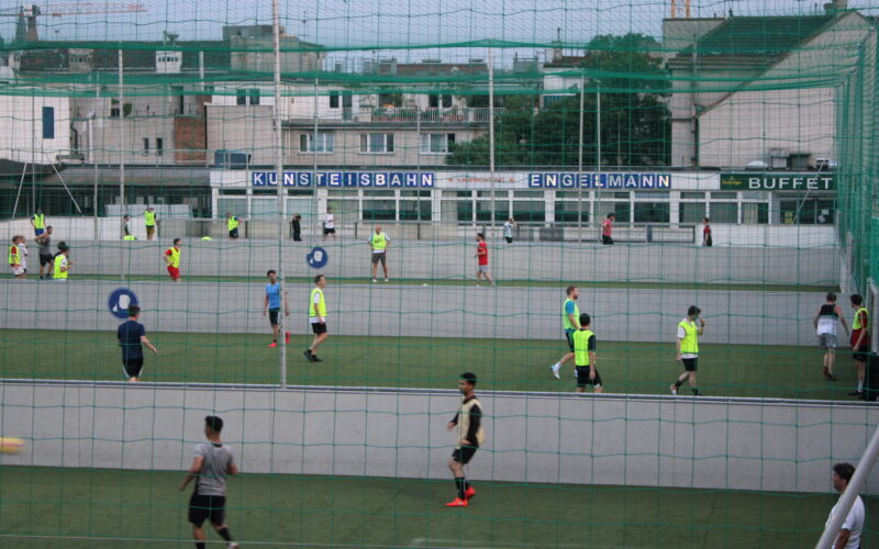 Mehrere Personen spielen Fußball auf einem mit Netzen eingefassten Feld im Freien, im Hintergrund sind Gebäude und ein Schild mit der Aufschrift "KUNSTEISBAHN ENGELMANN BUFFET" zu sehen.