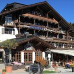 Ein großes Hotel im alpinen Stil mit Holzbalkonen und Sitzgelegenheiten im Freien, mit Bar und Restaurant unter blauem Himmel.