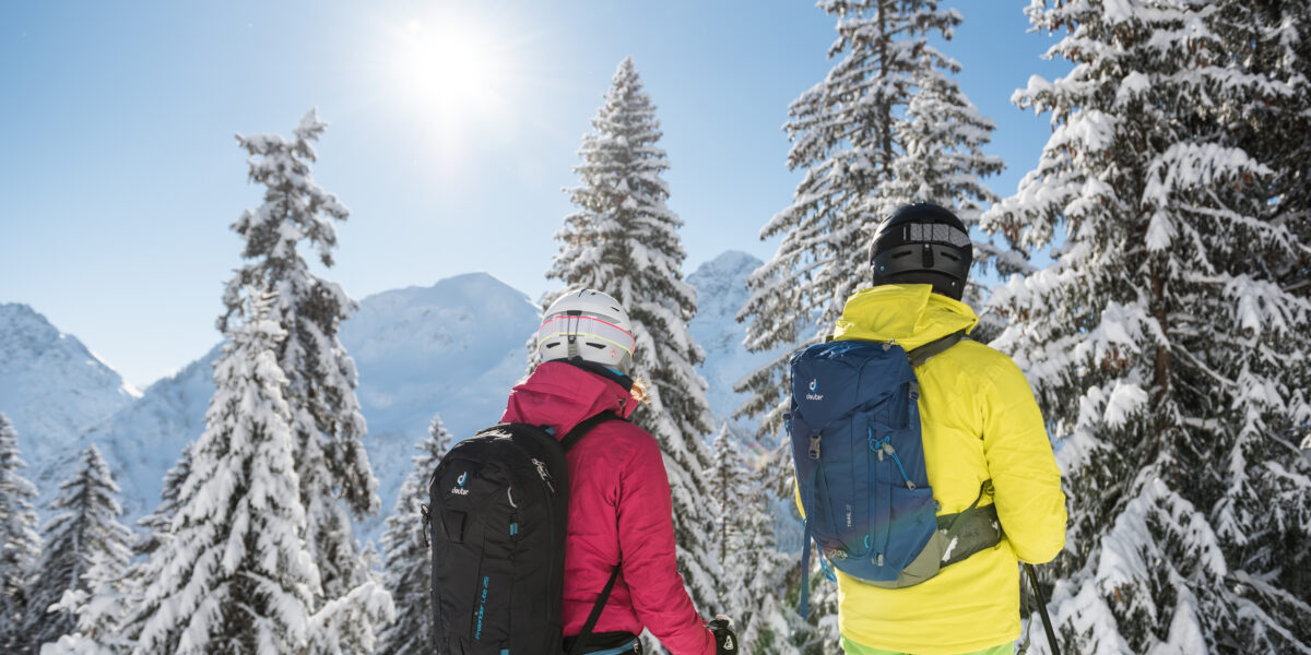 Zwei Menschen mit Helmen und Winterkleidung stehen zwischen schneebedeckten Bäumen und blicken auf eine Berglandschaft unter einem klaren blauen Himmel mit hellem Sonnenlicht.