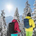 Zwei Menschen mit Helmen und Winterkleidung stehen zwischen schneebedeckten Bäumen und blicken auf eine Berglandschaft unter einem klaren blauen Himmel mit hellem Sonnenlicht.
