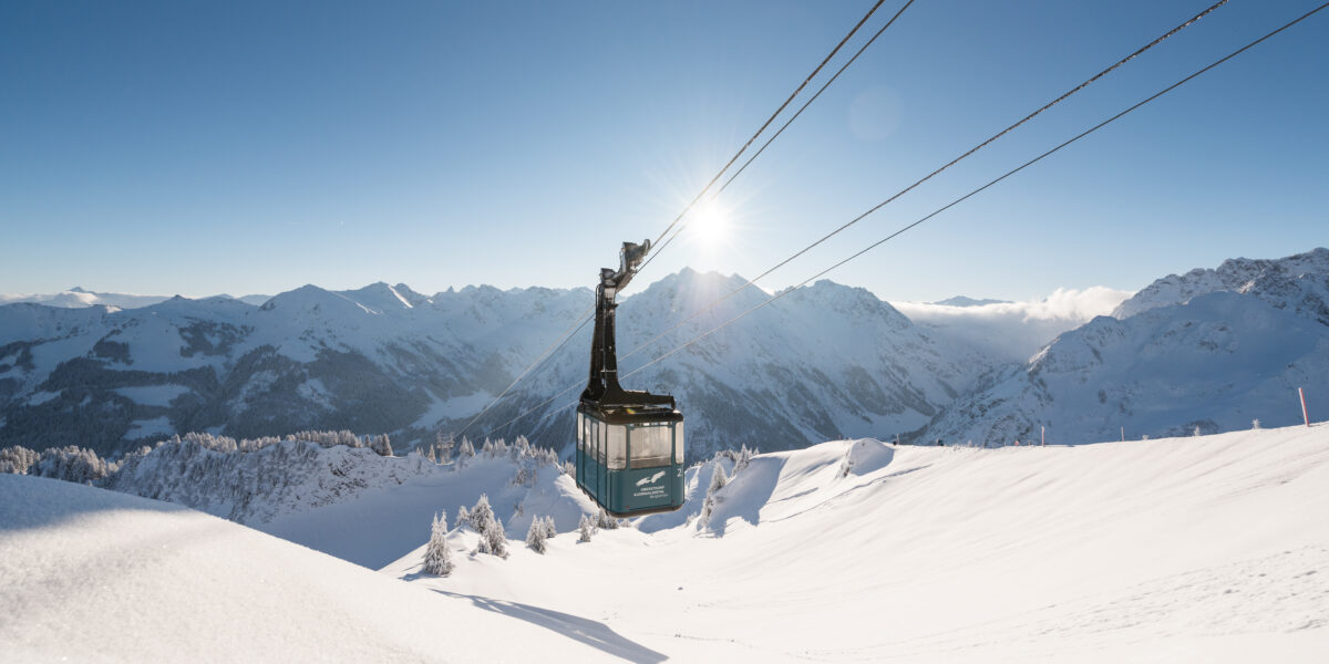 Eine Seilbahn fährt unter einem klaren blauen Himmel über schneebedeckte Berge, im Hintergrund scheint die Sonne.