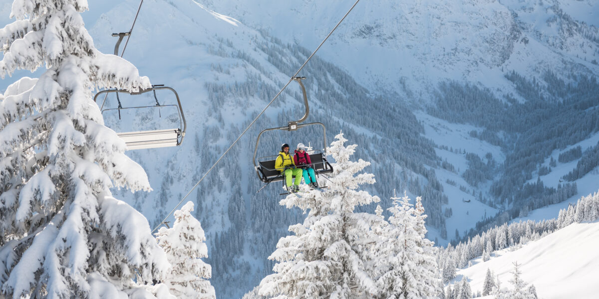 Zwei Personen in Skiausrüstung sitzen auf einem Skilift, der über schneebedeckte Bäume mit Bergen im Hintergrund hinauffährt.