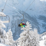 Zwei Personen in Skiausrüstung sitzen auf einem Skilift, der über schneebedeckte Bäume mit Bergen im Hintergrund hinauffährt.