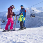 Drei Personen in Skiausrüstung stehen an einem klaren Tag auf einem schneebedeckten Hang mit Bergen und einer Gondelbahn im Hintergrund.