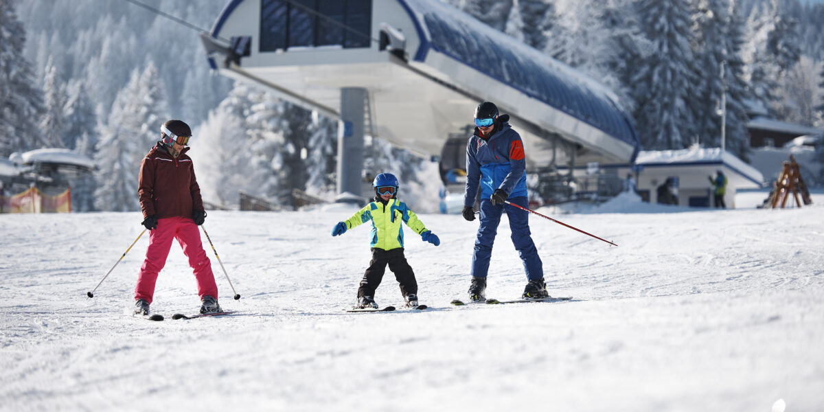 Drei Personen, darunter ein Kind, fahren auf einem verschneiten Hang in der Nähe einer Skiliftstation Ski, im Hintergrund sind schneebedeckte Bäume zu sehen.