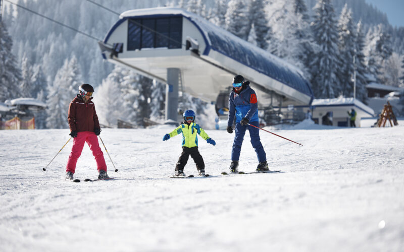 Drei Personen, darunter ein Kind, fahren auf einem verschneiten Hang in der Nähe einer Skiliftstation Ski, im Hintergrund sind schneebedeckte Bäume zu sehen.