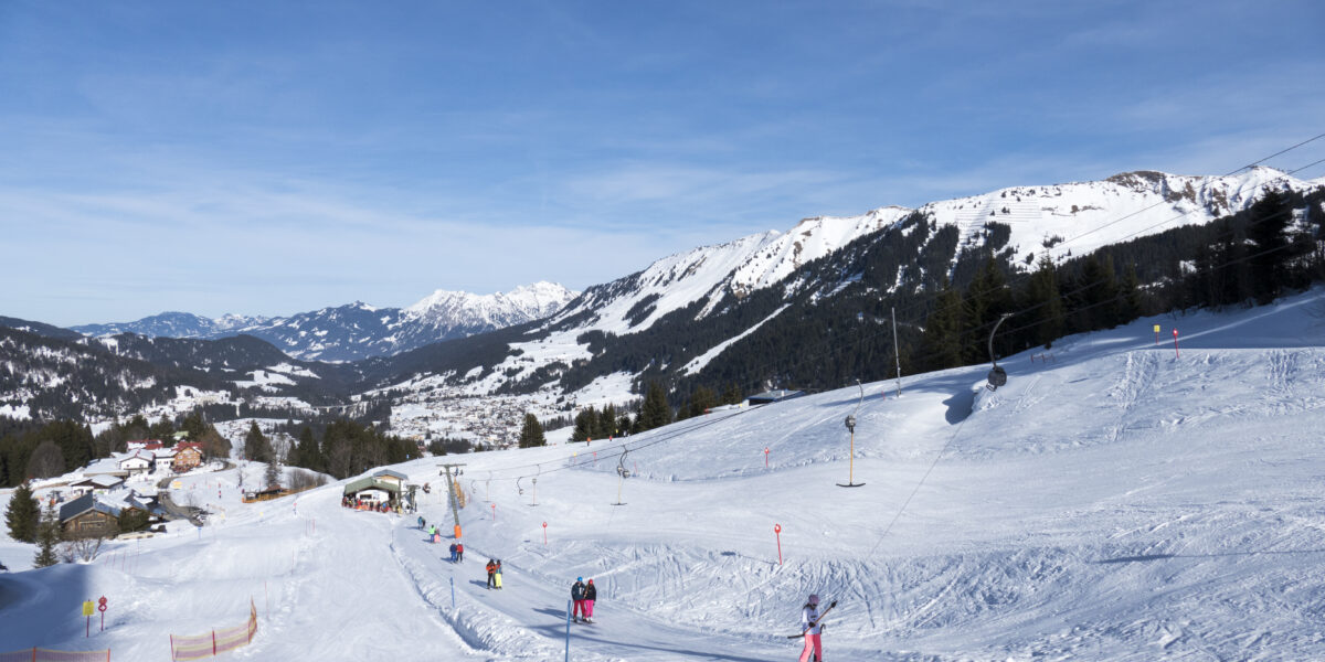 Skifahrer fahren einen schneebedeckten Hang in einem Skigebiet hinunter, im Hintergrund sind Berge und vereinzelte Bäume unter einem klaren blauen Himmel zu sehen.