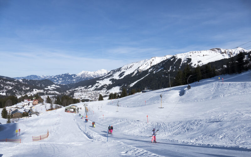 Skifahrer fahren einen schneebedeckten Hang in einem Skigebiet hinunter, im Hintergrund sind Berge und vereinzelte Bäume unter einem klaren blauen Himmel zu sehen.
