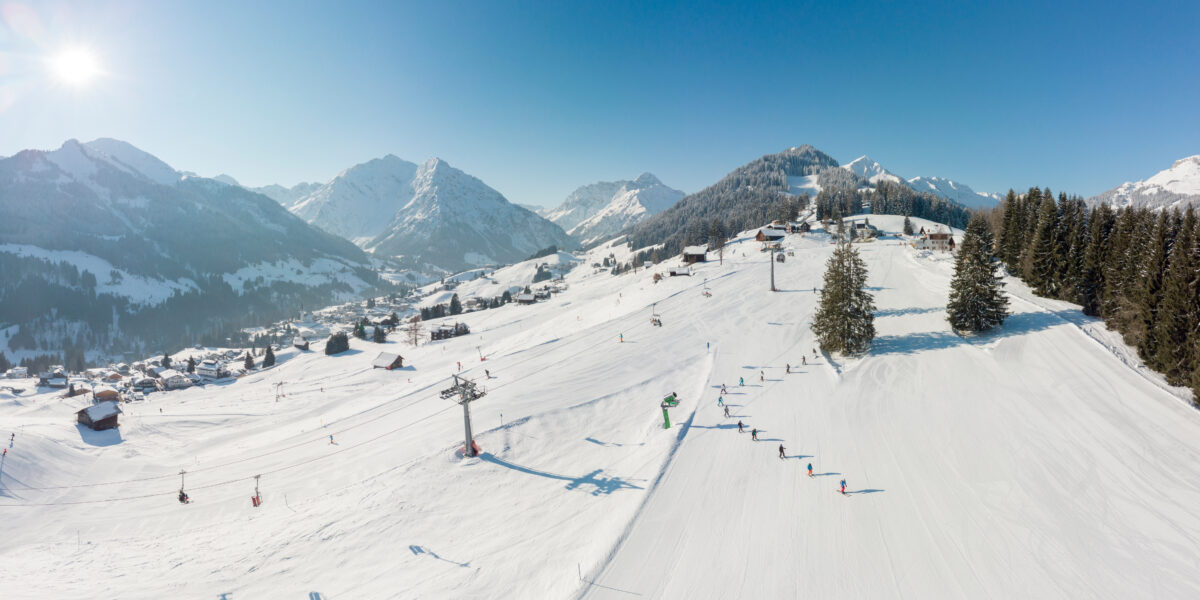 Luftaufnahme einer verschneiten Skipiste mit Skifahrern, Skilift und verstreuten Gebäuden; umgeben von Bergen und Kiefern unter einem klaren blauen Himmel.