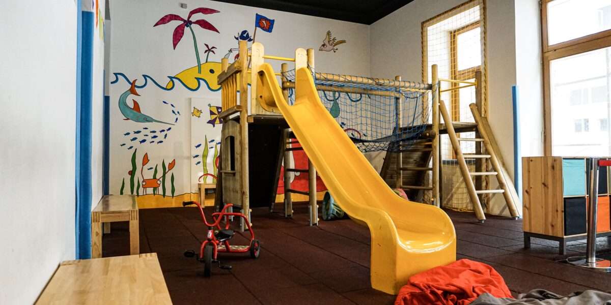 Innenspielzimmer mit einer gelben Rutsche, einem hölzernen Klettergerüst, Bänken, einem roten Dreirad und farbenfrohen Wandmalereien mit Meeresbewohnern und Pflanzen.