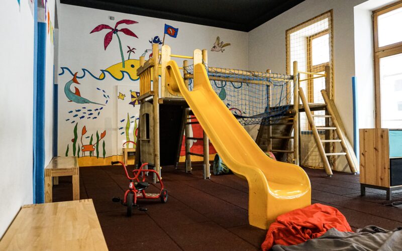 Innenspielzimmer mit einer gelben Rutsche, einem hölzernen Klettergerüst, Bänken, einem roten Dreirad und farbenfrohen Wandmalereien mit Meeresbewohnern und Pflanzen.