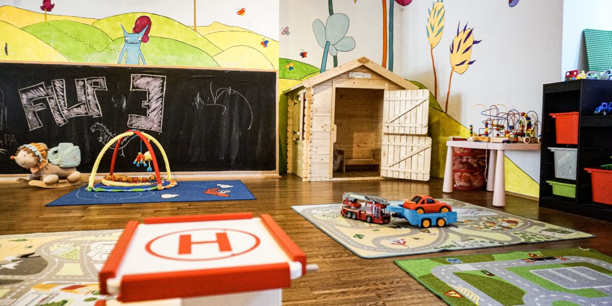 Buntes Kinderspielzimmer mit Wandmalerei, Holzspielhaus, Aktivitätsmatten, Spielzeugautos, Plüschtieren und Lernspielzeug in Regalen.