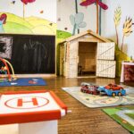 Buntes Kinderspielzimmer mit Wandmalerei, Holzspielhaus, Aktivitätsmatten, Spielzeugautos, Plüschtieren und Lernspielzeug in Regalen.