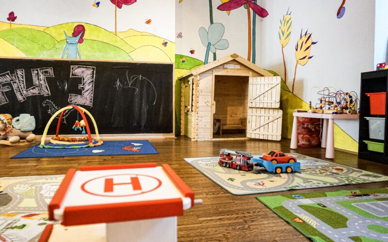 Buntes Kinderspielzimmer mit Wandmalerei, Holzspielhaus, Aktivitätsmatten, Spielzeugautos, Plüschtieren und Lernspielzeug in Regalen.