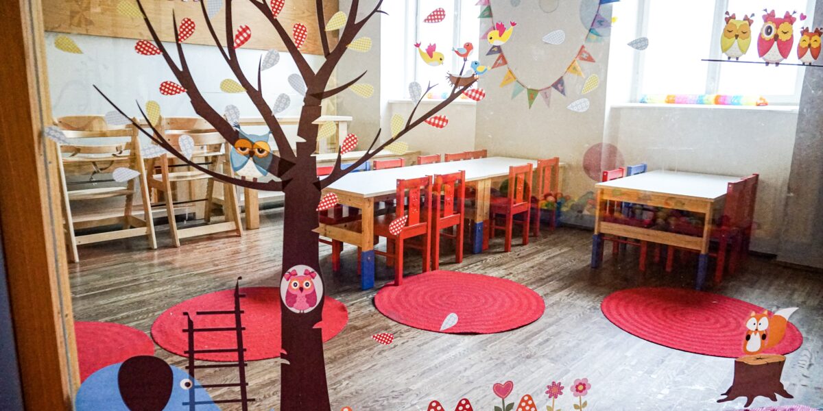 Ein hell dekoriertes Klassenzimmer mit kleinen Tischen, Stühlen, roten Teppichen und verspielten Wand- und Fensterbildern von Bäumen, Eulen und einer Elefantenleiter.