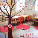 Ein hell dekoriertes Klassenzimmer mit kleinen Tischen, Stühlen, roten Teppichen und verspielten Wand- und Fensterbildern von Bäumen, Eulen und einer Elefantenleiter.