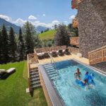 Zwei Personen entspannen sich in einem Außenpool neben einem Holzgebäude, mit Liegestühlen auf dem Rasen und Bergen im Hintergrund unter einem sonnigen Himmel.