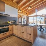 Moderne Küche mit Holzschränken, Steinsockel, freiliegenden Holzbalken, großen Fenstern und einem Essbereich mit Tageslicht und Bergblick.