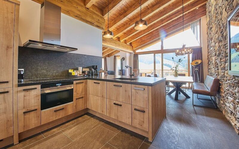 Moderne Küche mit Holzschränken, Steinsockel, freiliegenden Holzbalken, großen Fenstern und einem Essbereich mit Tageslicht und Bergblick.