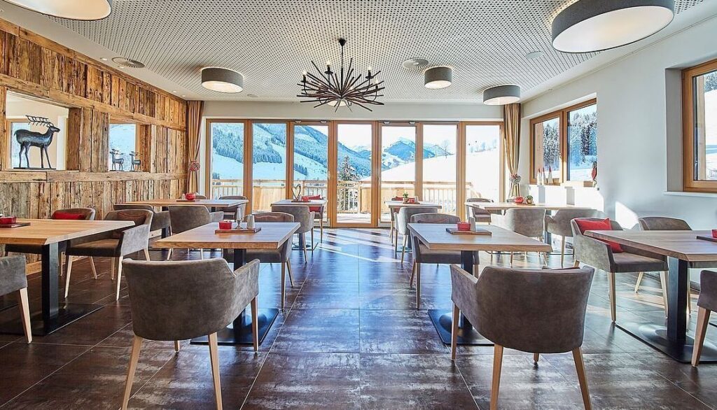 Ein moderner Essbereich mit Holztischen und grauen Stühlen, große Fenster mit Blick auf verschneite Berge und eine dekorative Holzwand mit einer Rentierfigur.