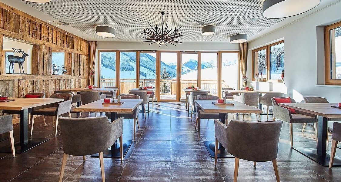 Moderner Essbereich mit Holztischen und grauen Stühlen, großen Fenstern mit Blick auf die Berge, Akzentwand aus Holz und modernen Deckenleuchten.