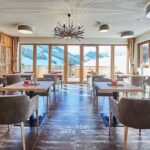 Moderner Essbereich mit Holztischen und grauen Stühlen, großen Fenstern mit Blick auf die Berge, Akzentwand aus Holz und modernen Deckenleuchten.