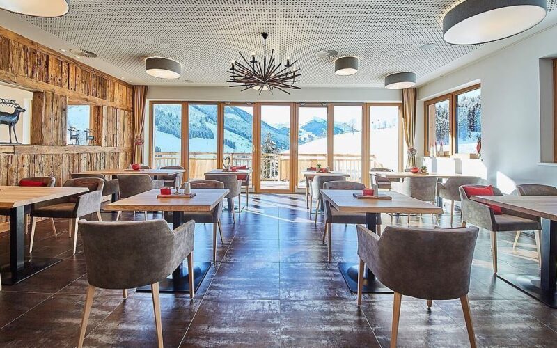 Moderner Essbereich mit Holztischen und grauen Stühlen, großen Fenstern mit Blick auf die Berge, Akzentwand aus Holz und modernen Deckenleuchten.