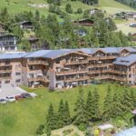 Ein großes, hölzernes Gebäude im Chalet-Stil mit Balkonen steht auf einem Hügel, umgeben von Gras, Bäumen und anderen ähnlichen Häusern in einer bergigen Gegend.