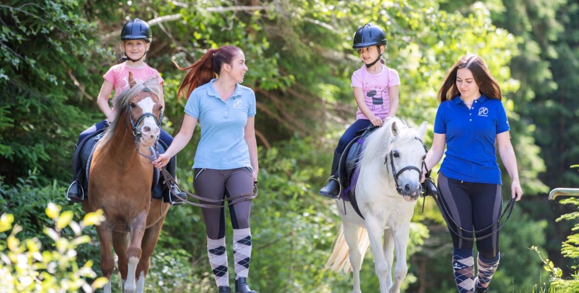 Zwei Mädchen reiten auf Ponys, während zwei Frauen an den Zügeln neben ihnen auf einem Waldweg gehen. Alle tragen Helme oder Reitkleidung.