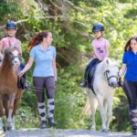 Zwei Mädchen reiten auf Ponys, während zwei Frauen an den Zügeln neben ihnen auf einem Waldweg gehen. Alle tragen Helme oder Reitkleidung.