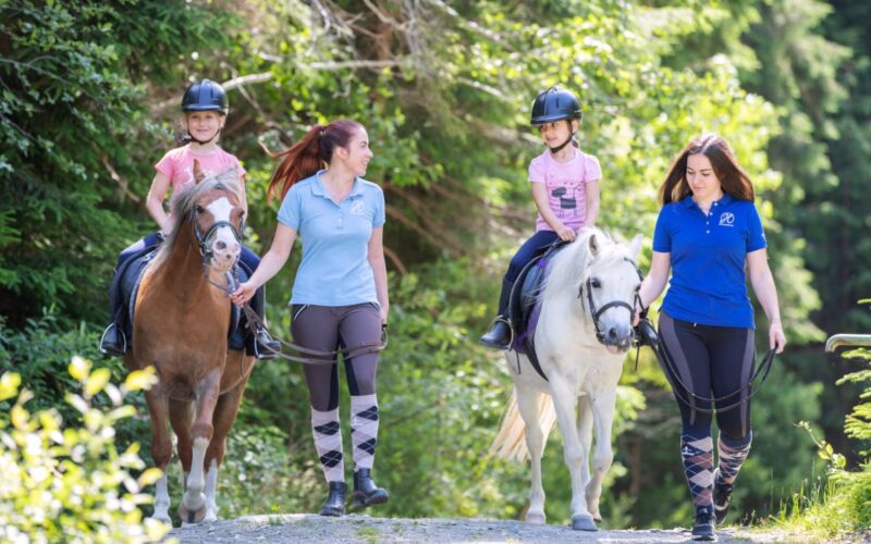 Zwei Mädchen reiten auf Ponys, während zwei Frauen an den Zügeln neben ihnen auf einem Waldweg gehen. Alle tragen Helme oder Reitkleidung.