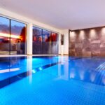 Hallenbad mit blauem Wasser, großen, bodentiefen Fenstern, Steinmauer und eingelassener Beleuchtung, mit Topfpflanzen in der Ecke.