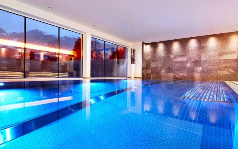 Hallenbad mit blauem Wasser, großen, bodentiefen Fenstern, Steinmauer und eingelassener Beleuchtung, mit Topfpflanzen in der Ecke.