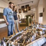 Zwei Personen stehen in einem Museum und betrachten ein detailliertes, maßstabsgetreues Modell einer historischen Stadt; an den Wänden hinter ihnen sind Gemälde und Exponate ausgestellt.