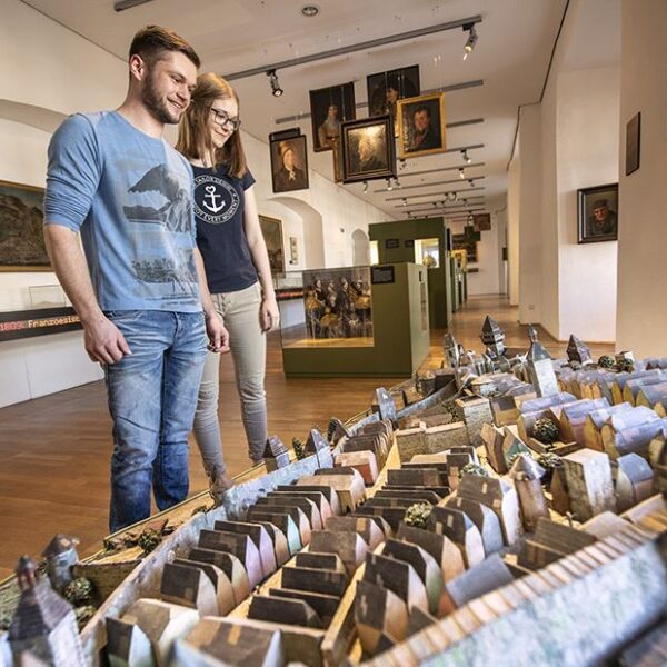 Zwei Personen stehen in einem Museum und betrachten ein detailliertes, maßstabsgetreues Modell einer historischen Stadt; an den Wänden hinter ihnen sind Gemälde und Exponate ausgestellt.