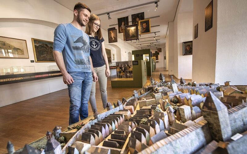 Zwei Personen stehen in einem Museum und betrachten ein detailliertes, maßstabsgetreues Modell einer historischen Stadt; an den Wänden hinter ihnen sind Gemälde und Exponate ausgestellt.