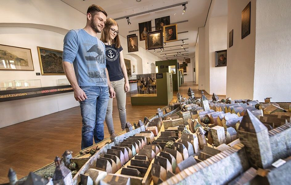 Zwei Personen stehen in einem Museum und betrachten ein detailliertes, maßstabsgetreues Modell einer historischen Stadt; an den Wänden hinter ihnen sind Gemälde und Exponate ausgestellt.