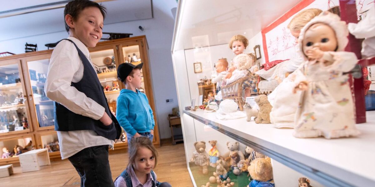 Drei Kinder betrachten eine Vitrine mit alten Puppen und Teddybären in einer Museumsumgebung.