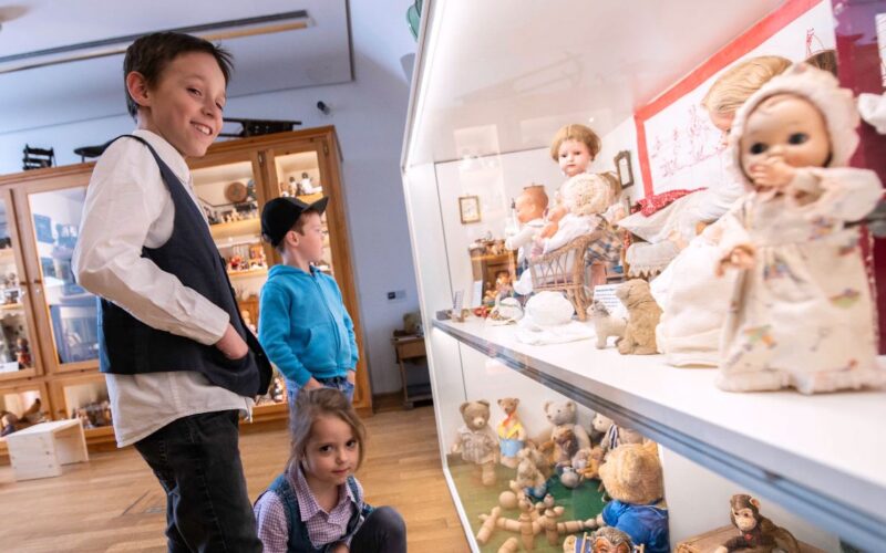 Drei Kinder betrachten eine Vitrine mit alten Puppen und Teddybären in einer Museumsumgebung.