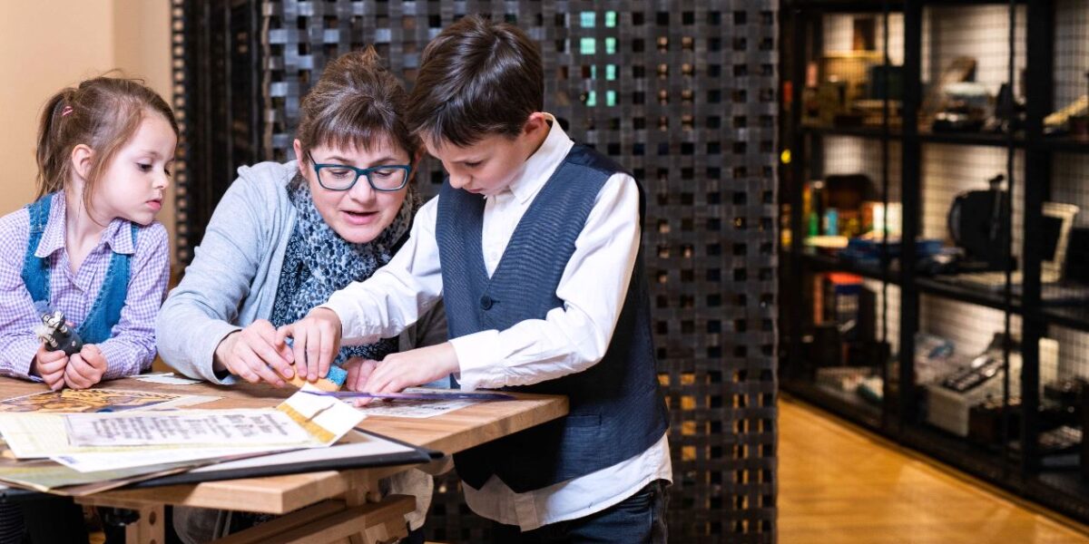 Ein Erwachsener und zwei Kinder arbeiten an einem Tisch in einem Museum oder Lernzentrum an einer pädagogischen Aktivität, mit Ausstellungsregalen im Hintergrund.