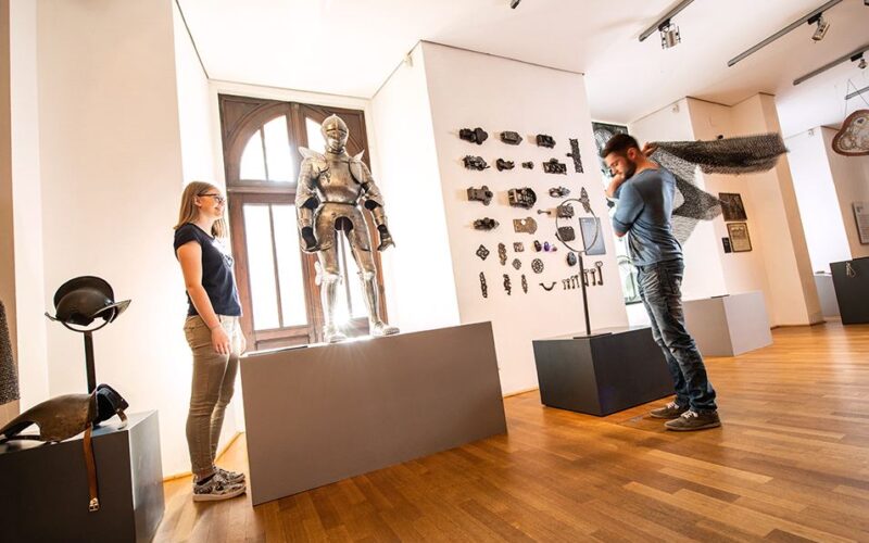 Zwei Personen betrachten und interagieren mit mittelalterlichen Rüstungen und Waffen in einem Ausstellungsraum mit Holzboden und weißen Wänden.