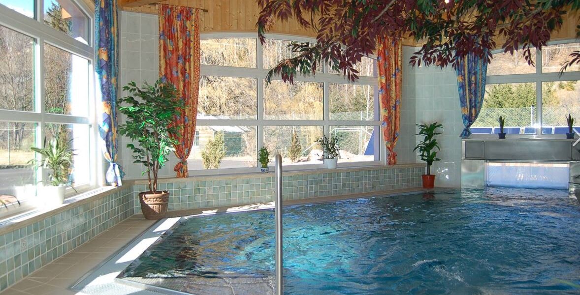 Hallenbad mit gefliestem Rand und mehreren Topfpflanzen, umgeben von großen Fenstern mit bunten Vorhängen und einer Holzdecke.