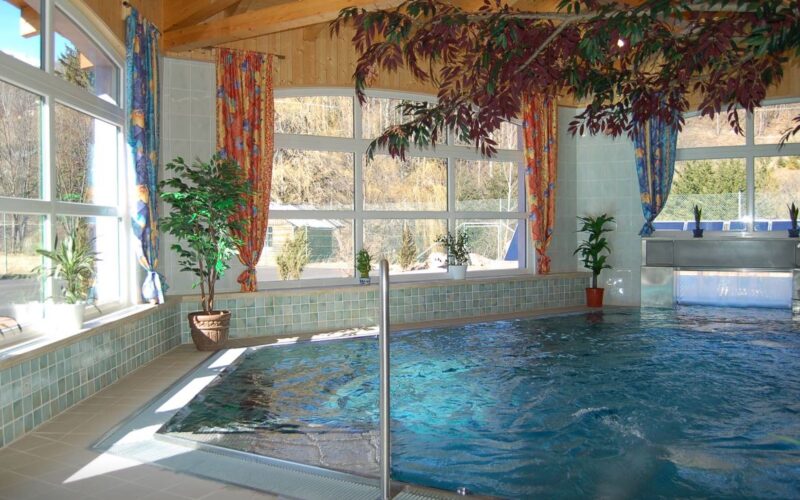 Hallenbad mit gefliestem Rand und mehreren Topfpflanzen, umgeben von großen Fenstern mit bunten Vorhängen und einer Holzdecke.