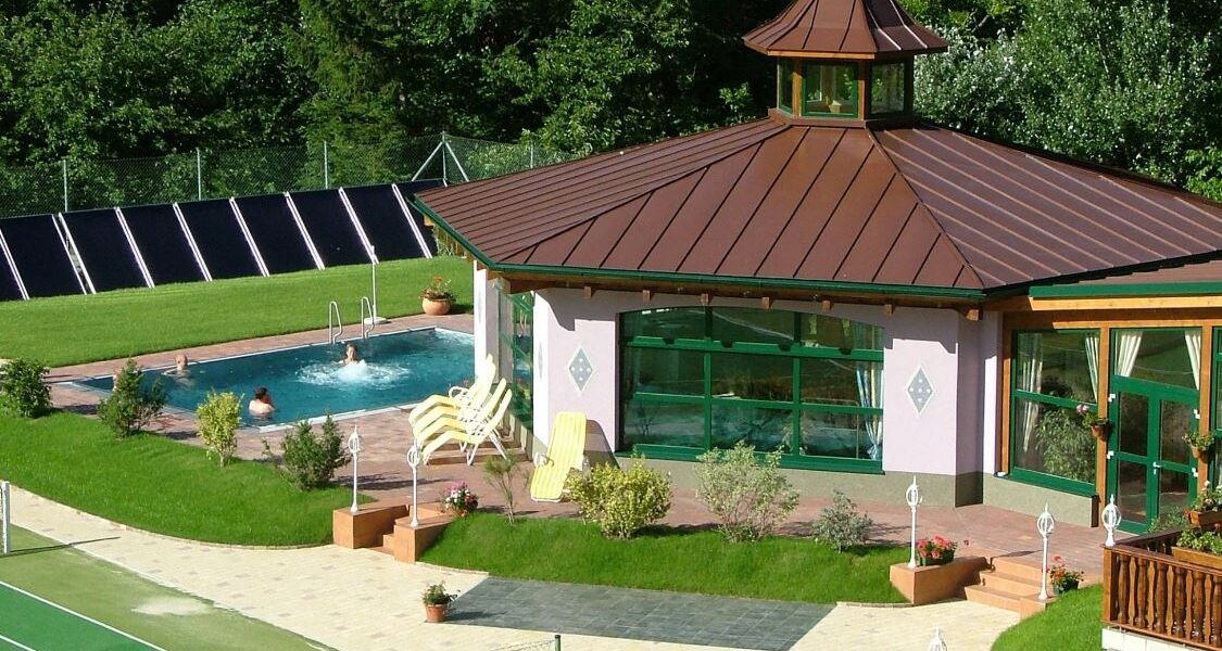 Ein kleiner Swimmingpool mit Liegestühlen befindet sich neben einem Pavillon mit großen Fenstern und einem braunen Dach, umgeben von Grünanlagen und einem Tennisplatz.