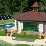 Ein kleiner Swimmingpool mit Liegestühlen befindet sich neben einem Pavillon mit großen Fenstern und einem braunen Dach, umgeben von Grünanlagen und einem Tennisplatz.